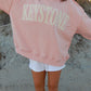 Person wearing a pink sweatshirt with 'Keystone' printed on it, standing on a sandy surface.