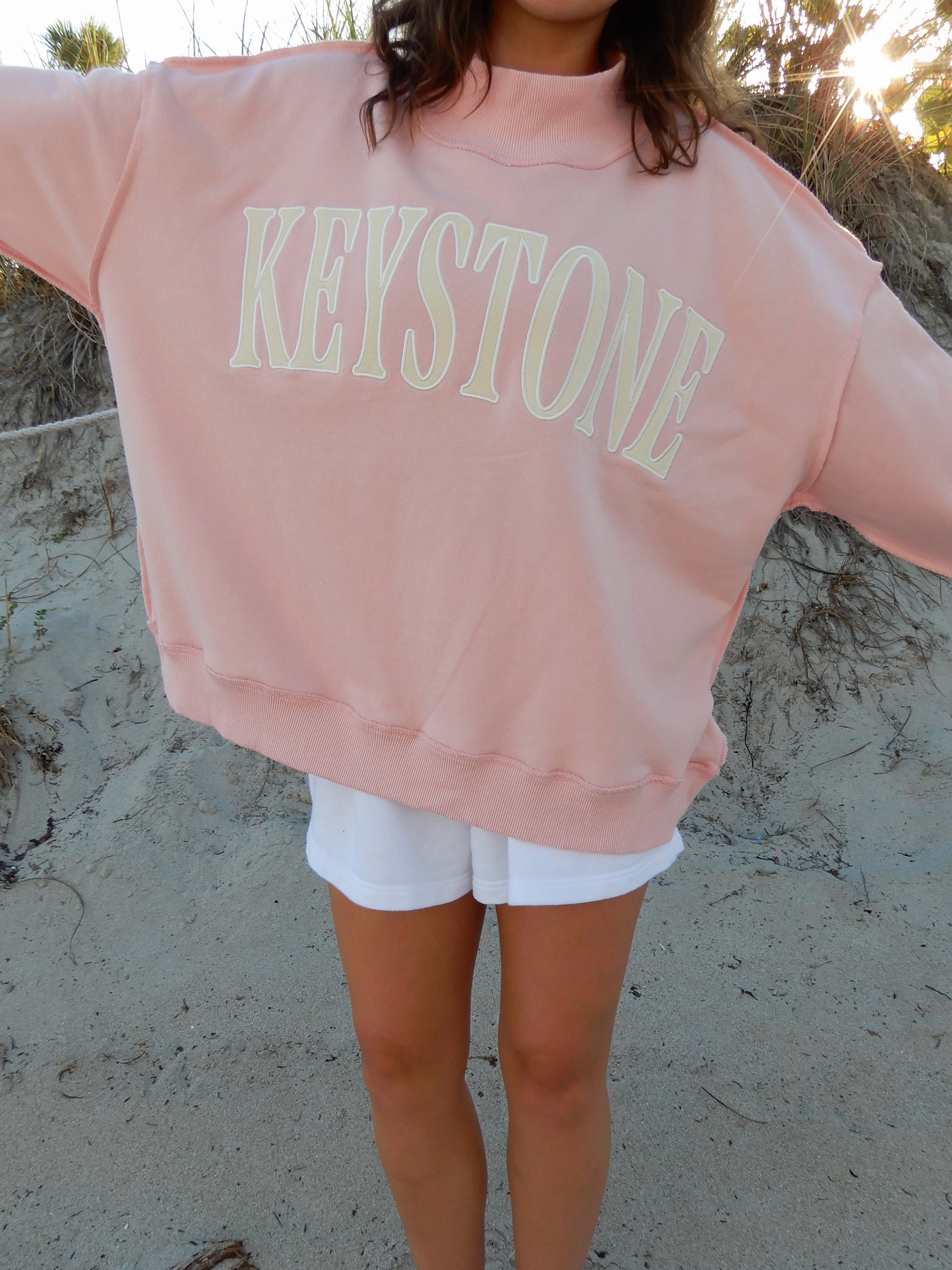 Person wearing a pink sweatshirt with 'Keystone' printed on it, standing on a sandy surface.