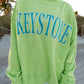Person wearing a bright green sweatshirt with 'Keystone' printed on it, standing on a beach.