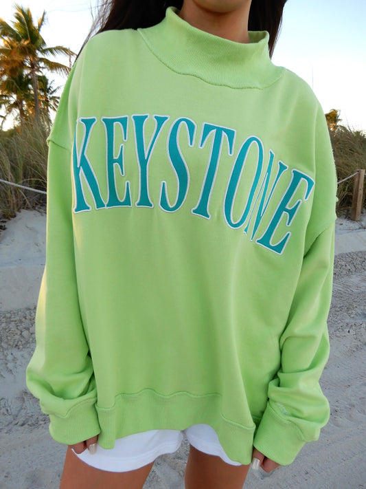 Person wearing a bright green sweatshirt with 'Keystone' printed on it, standing on a beach.