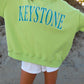Person wearing a bright green sweatshirt with 'Keystone' printed on it, standing on sandy ground.