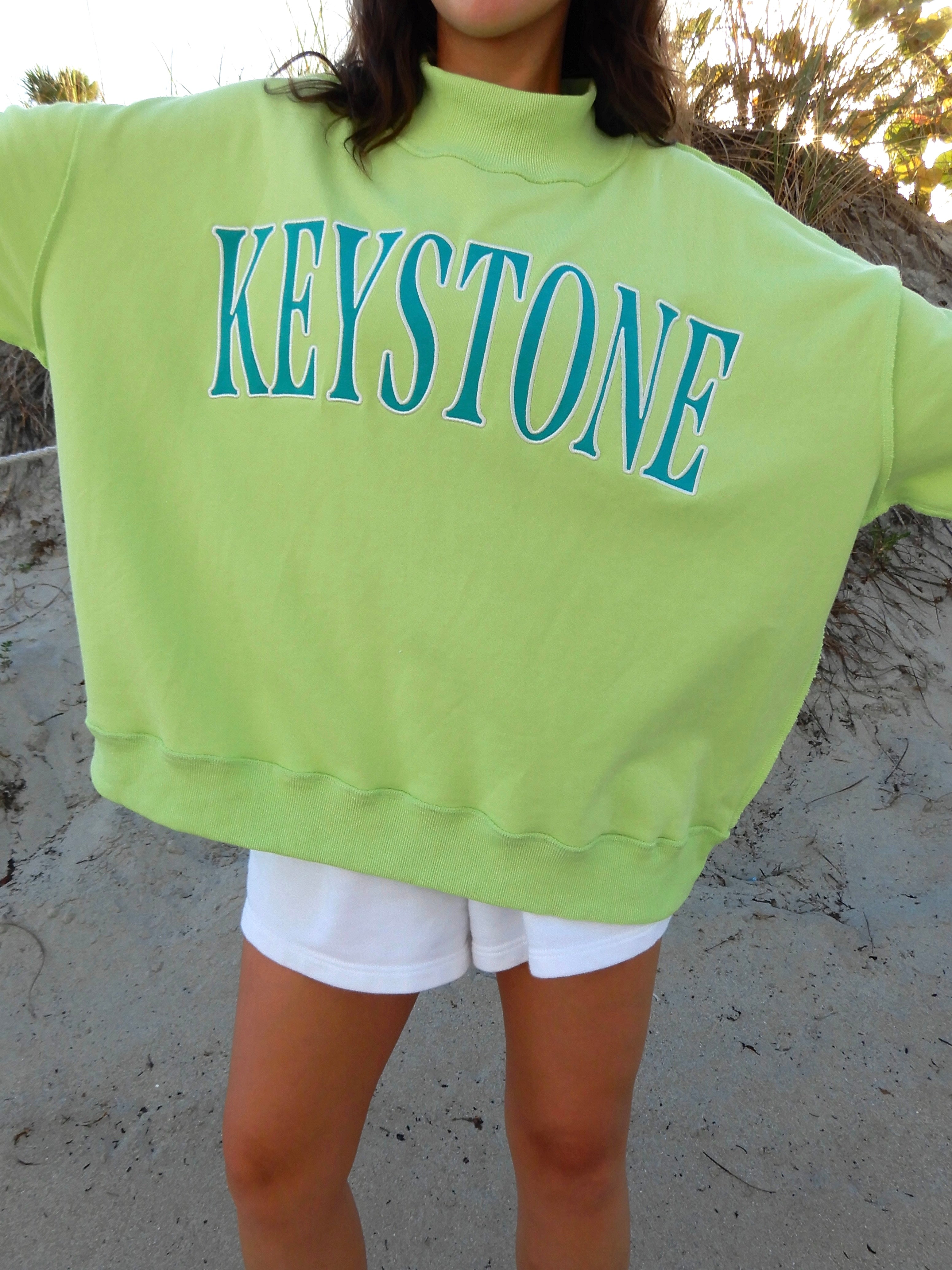 Person wearing a bright green sweatshirt with 'Keystone' printed on it, standing on sandy ground.