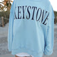 Person wearing a light blue sweatshirt with 'Keystone' text on a beach.