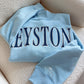 Light blue sweatshirt with 'KEYSTONE' printed on it, laid flat on a beige surface.