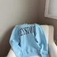 Light blue sweatshirt with text on a beige chair against a neutral wall.