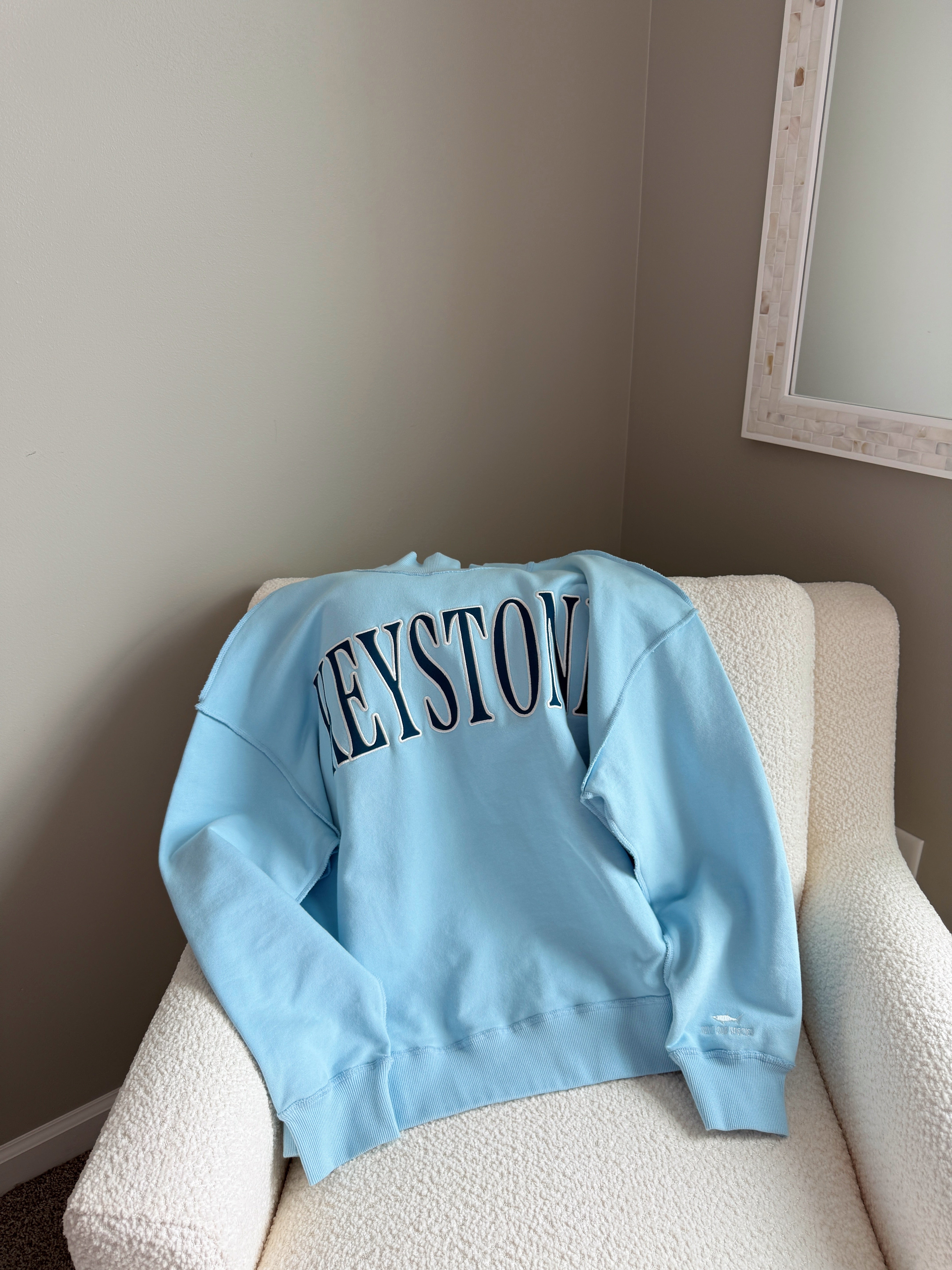 Light blue sweatshirt with text on a beige chair against a neutral wall.