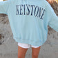 Person wearing a blue sweatshirt with dark 'KEYSTONE' text on a sandy surface