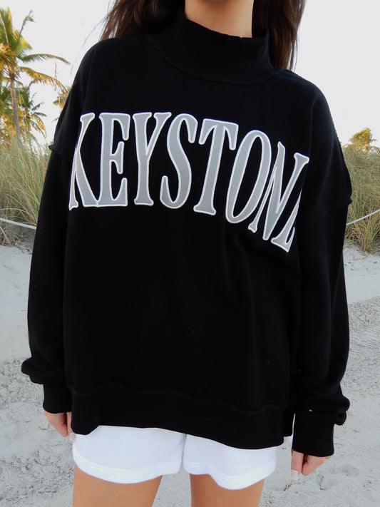 Person wearing a black sweatshirt with 'KESTONE' printed on it, standing on a sandy beach.