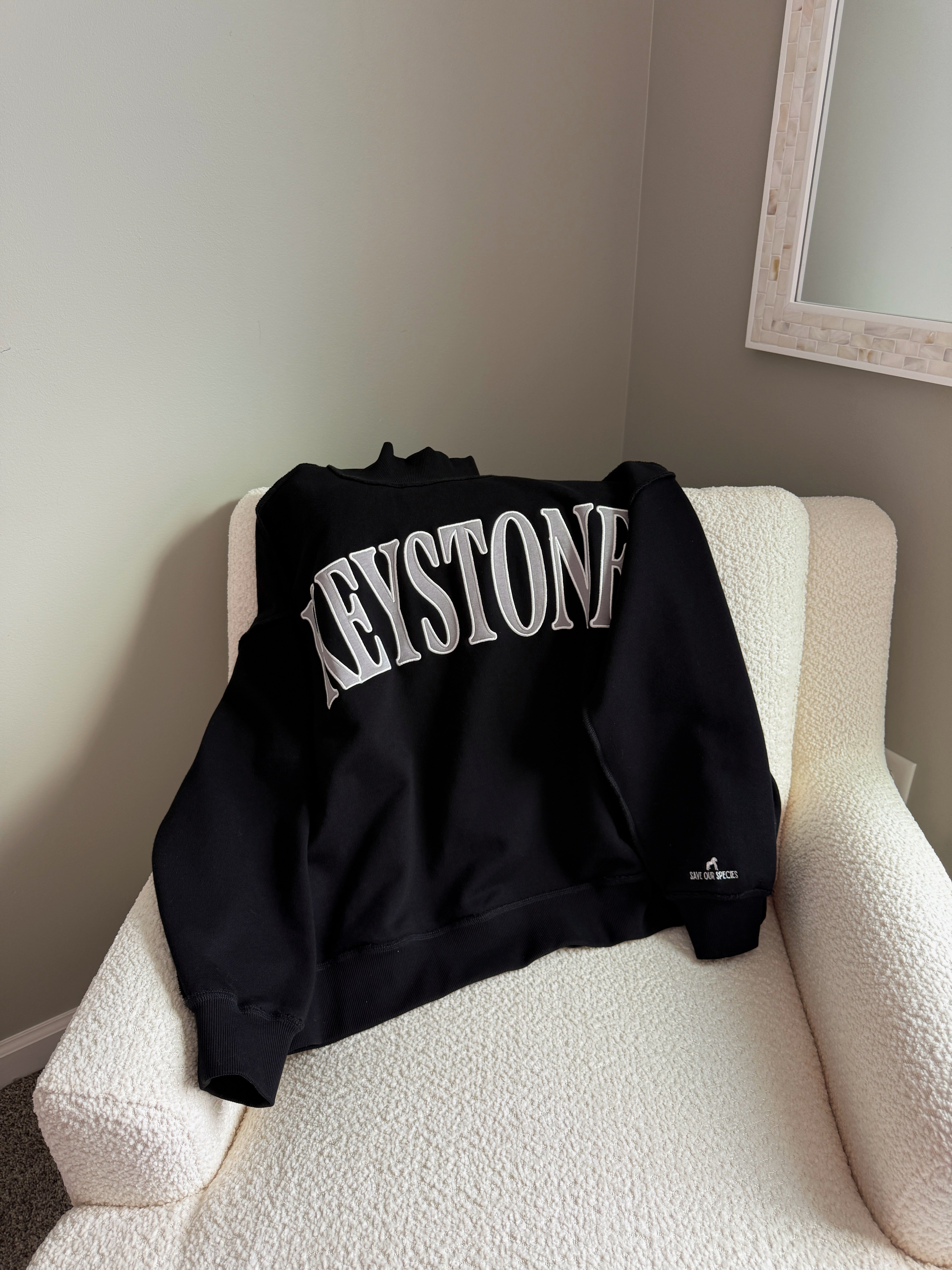 Black sweatshirt with 'Keystone' text on a white chair against a gray wall.