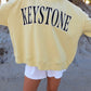 Person wearing a yellow sweatshirt with 'Keystone' printed on it, standing on sandy ground.
