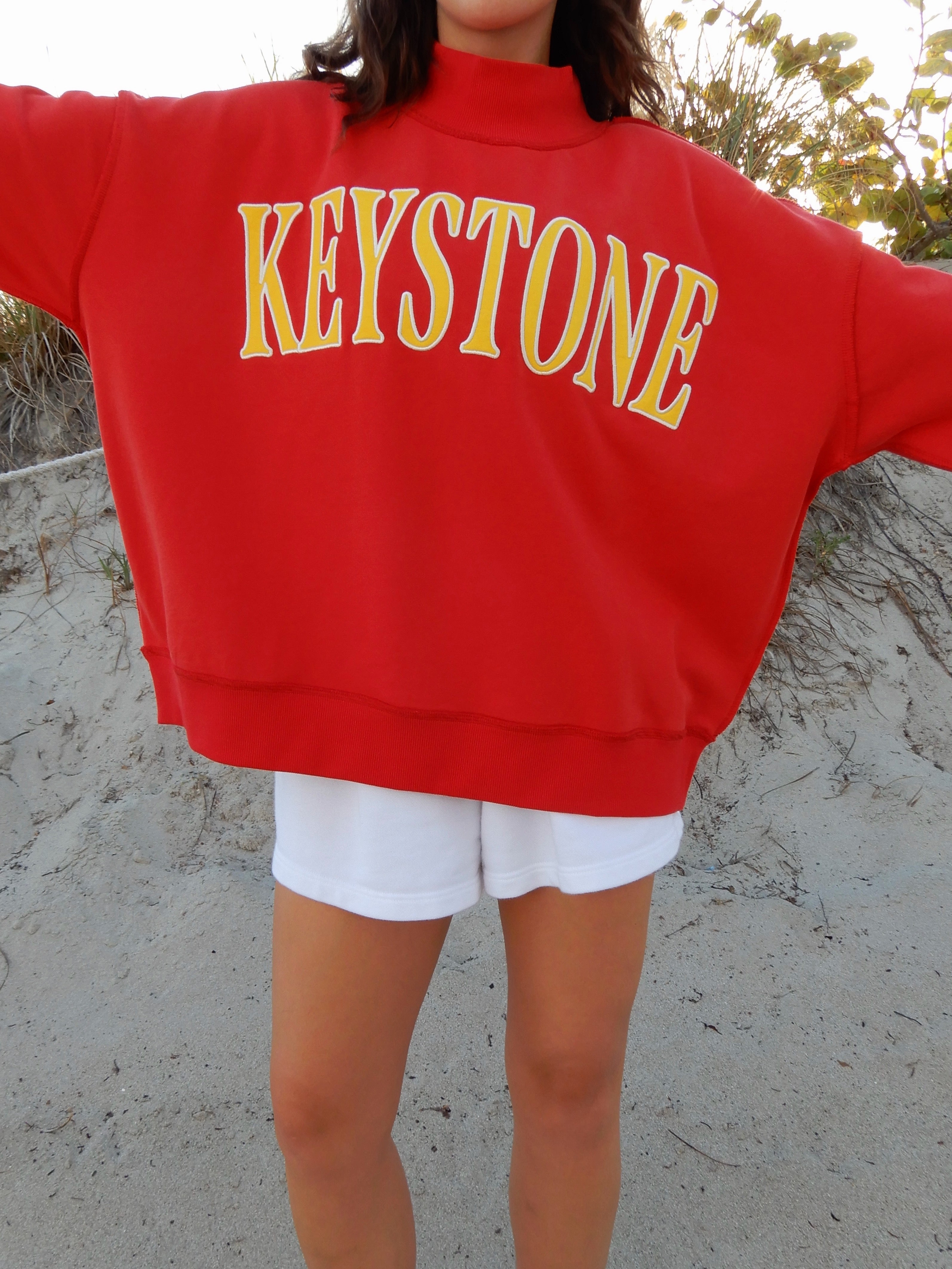 Person wearing a red sweatshirt with 'Keystone' printed in yellow, standing on a sandy surface.