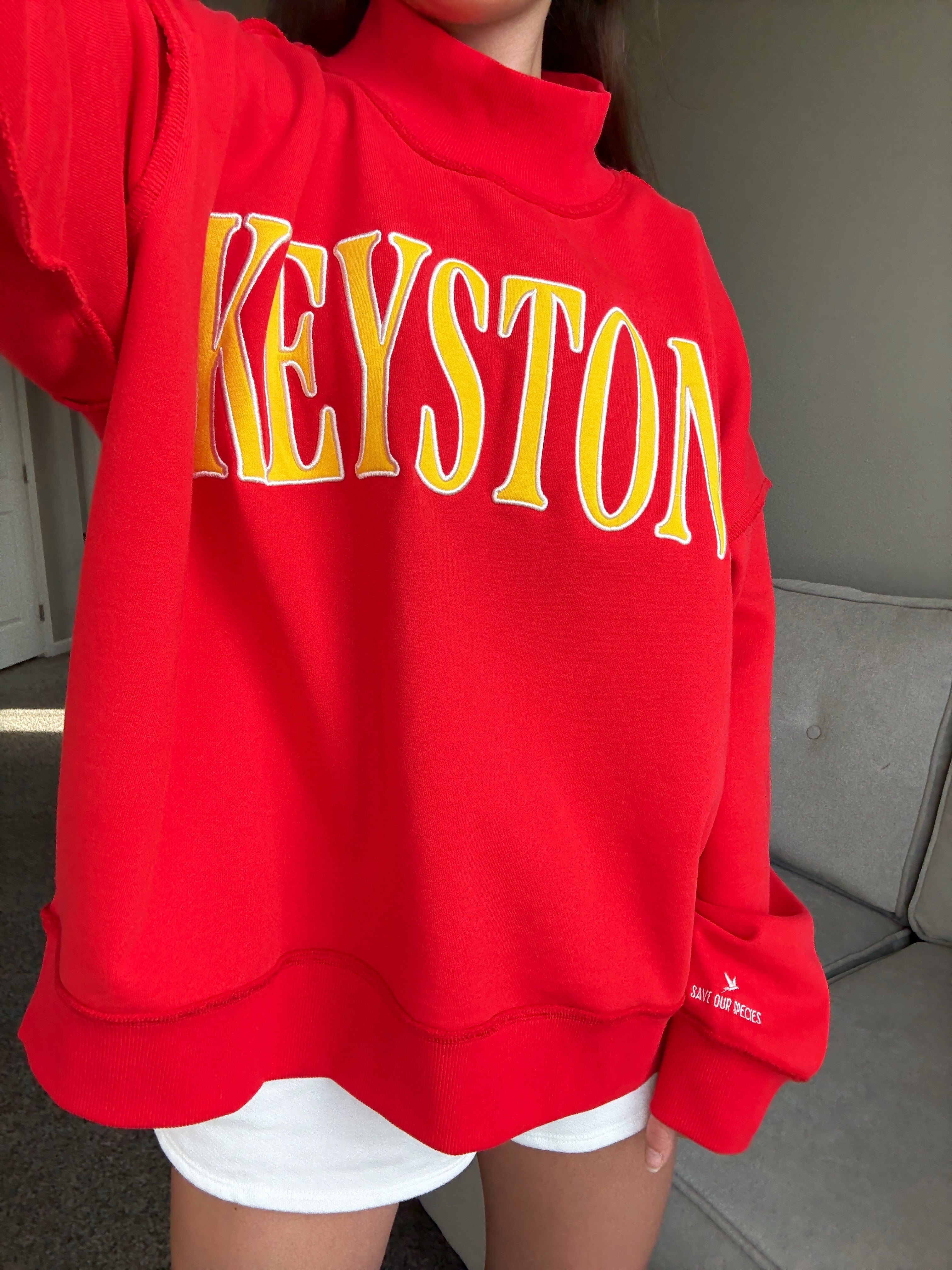 Red sweatshirt with yellow 'Keystone' text on a model in front of a couch
